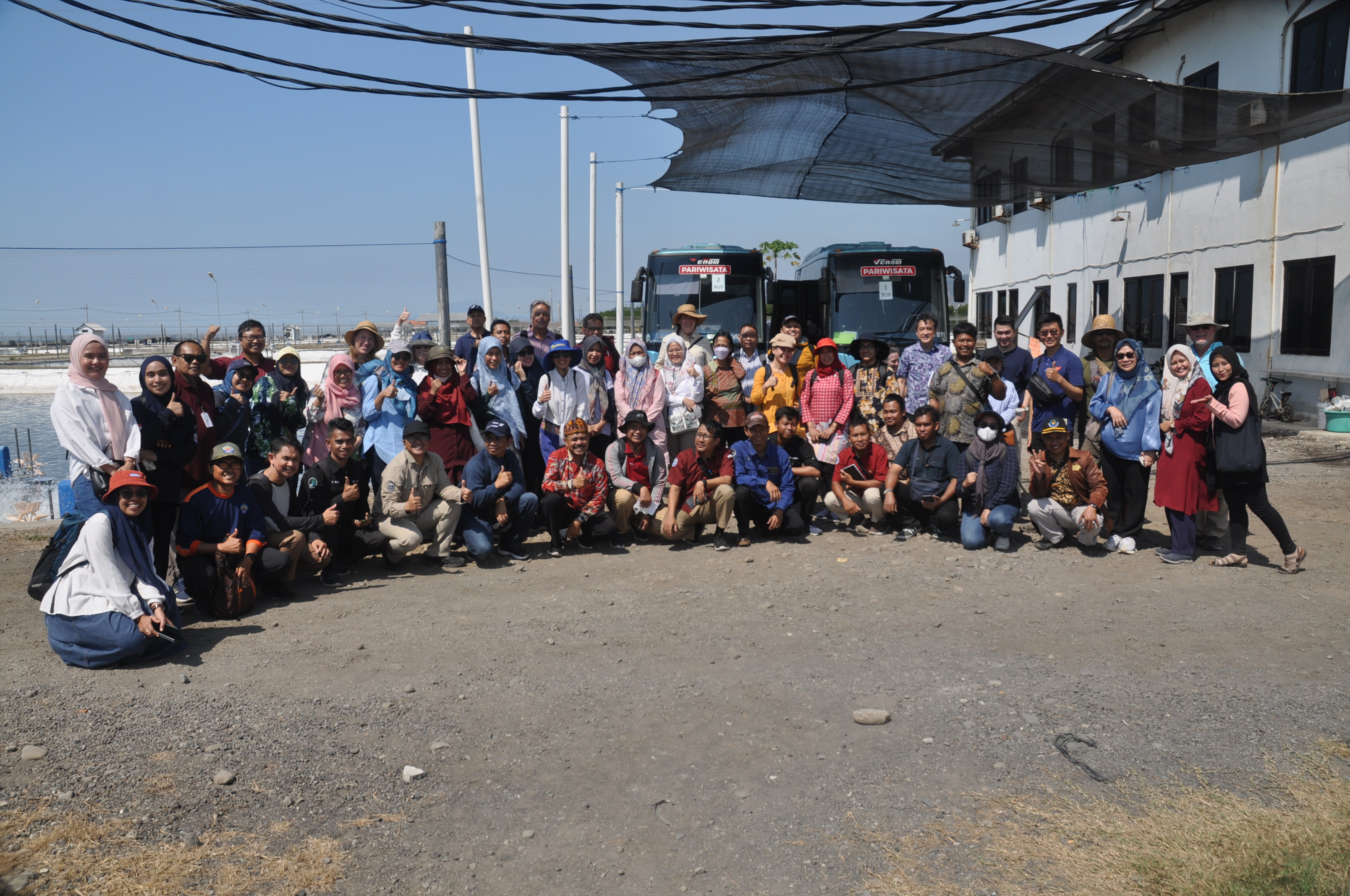 Group photo of participants in front of two tour buses, under shade netting, at the shrimp farm.