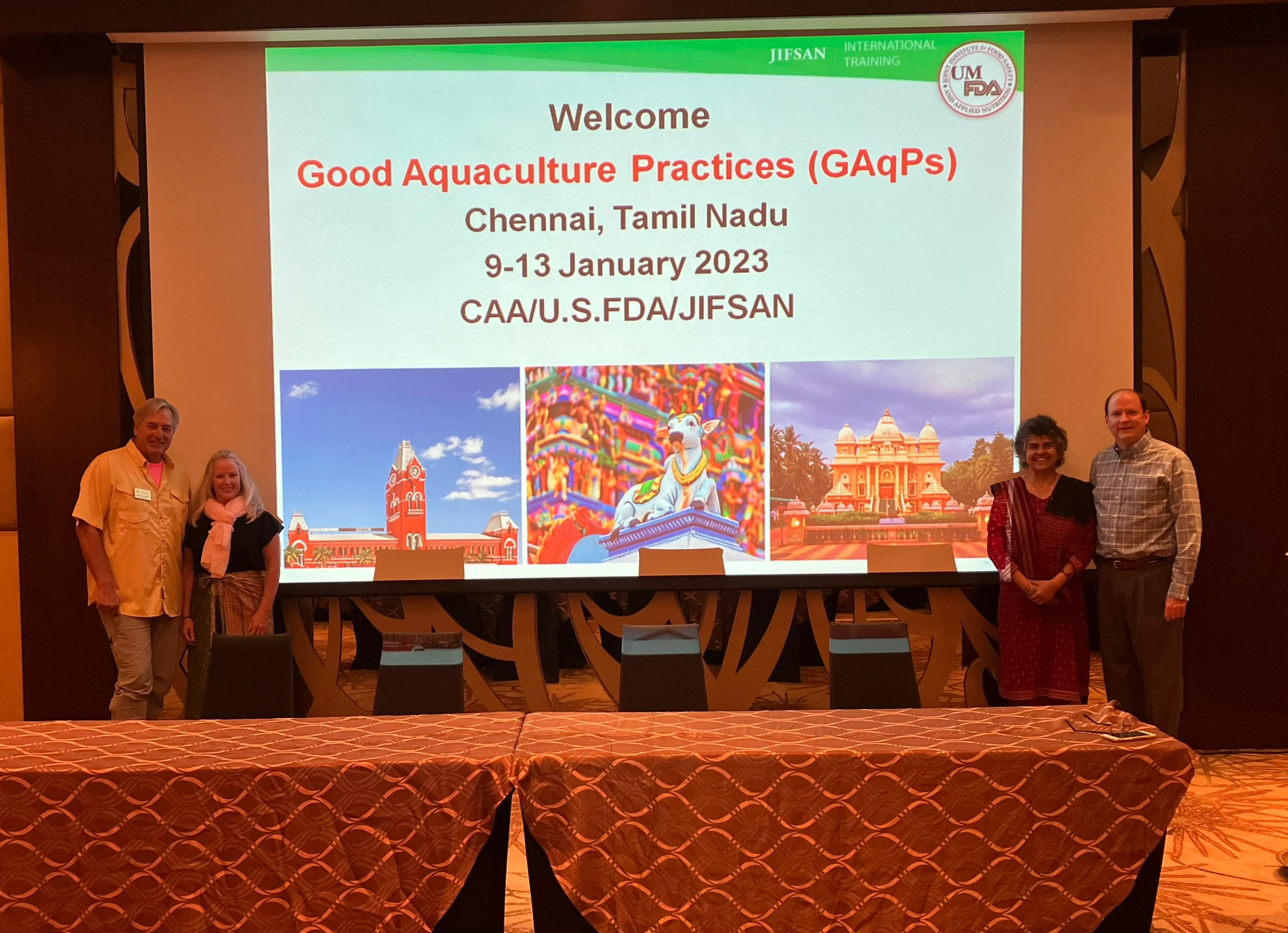 Four conference participants stand in front of a projected slide that reads 'Welcome; Good Aquacultural Practices (GAqP); Chennai, Tamil Nadu' with images of temples and other sites around the city of Chennai.